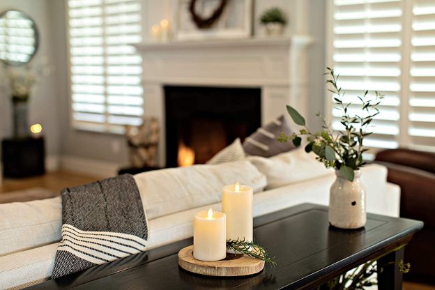 2 ivory flameless LED Candles displayed on a table next to a couch. Ferns and a plant are decorated around the candles.